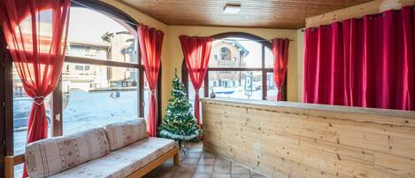 A wooden interior with a bench, a Christmas tree, and large windows with red curtains.
