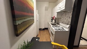 Una cocina compacta con gabinetes blancos, una estufa a gas y un lavamanos. Hay un cuadro en la pared y una planta sobre la mesa.
