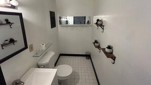 Un baño pequeño con inodoro, lavamanos y espejo. Hay repisas con plantas en macetas y una ventana sobre el inodoro.