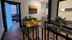 Cocina moderna con estufa a gas, armarios blancos y encimera negra. Hay una zona de comedor con sillas amarillas y una mesa negra. Un cuadro cuelga de la pared y hay una planta en la encimera.