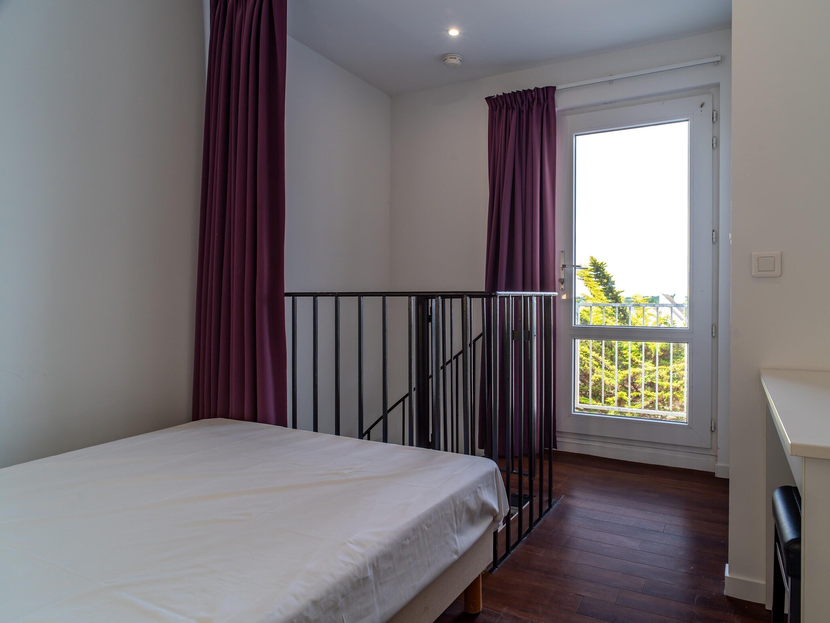 A bedroom with a bed, a window with curtains, a balcony, and a door.