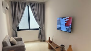 A modern living room with a grey sofa, a wooden coffee table, a TV mounted on the wall, and a large window with curtains.