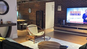 Een moderne woonkamer met een televisie waarop 'Inside Story' te zien is, een bank, een houten bijzettafeltje en een rieten fauteuil.