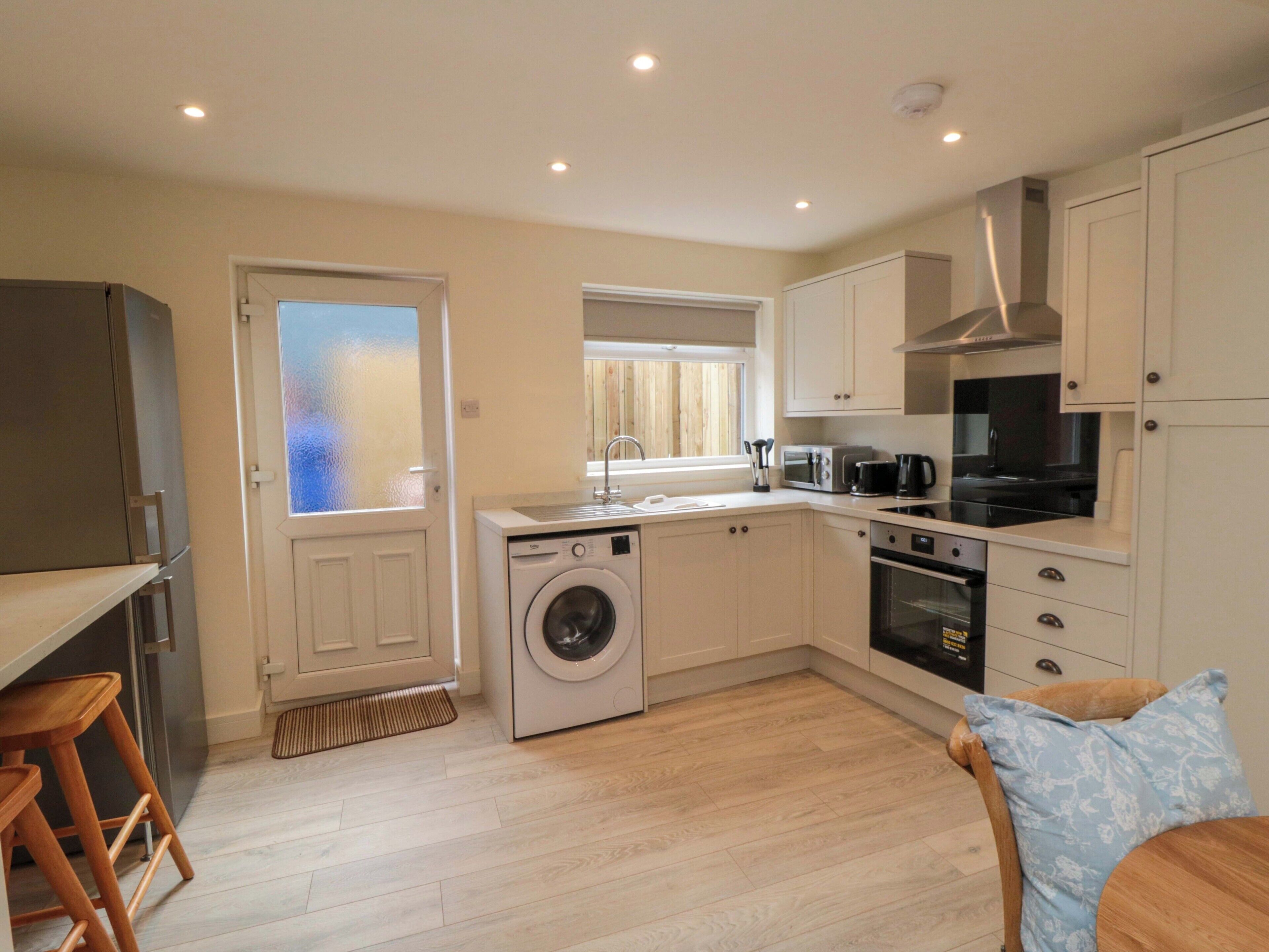 A compact kitchen with a washing machine, microwave, oven, and a dining area with a chair and table.