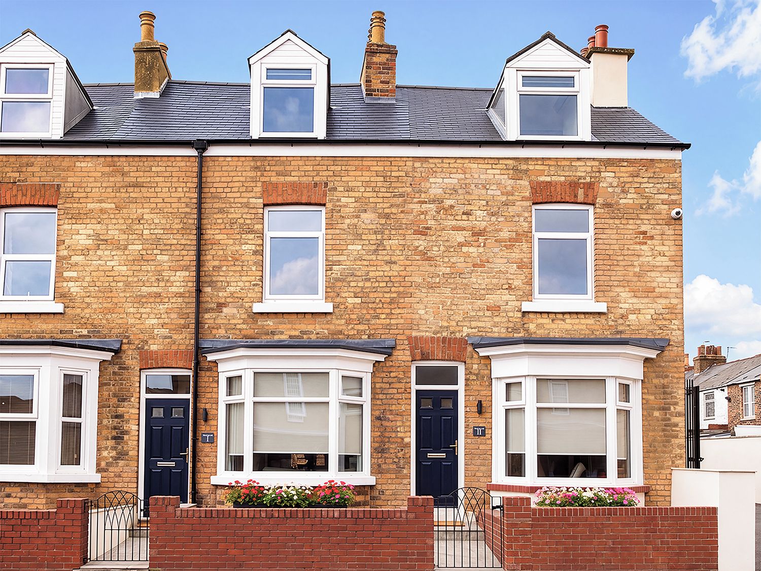 A two-story terraced house with a brick facade, white-framed windows, and a front door with a small garden area with flowers.