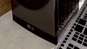Une machine à laver LG noire à chargement frontal, dans une pièce aux murs recouverts de carreaux blancs et au sol marron.