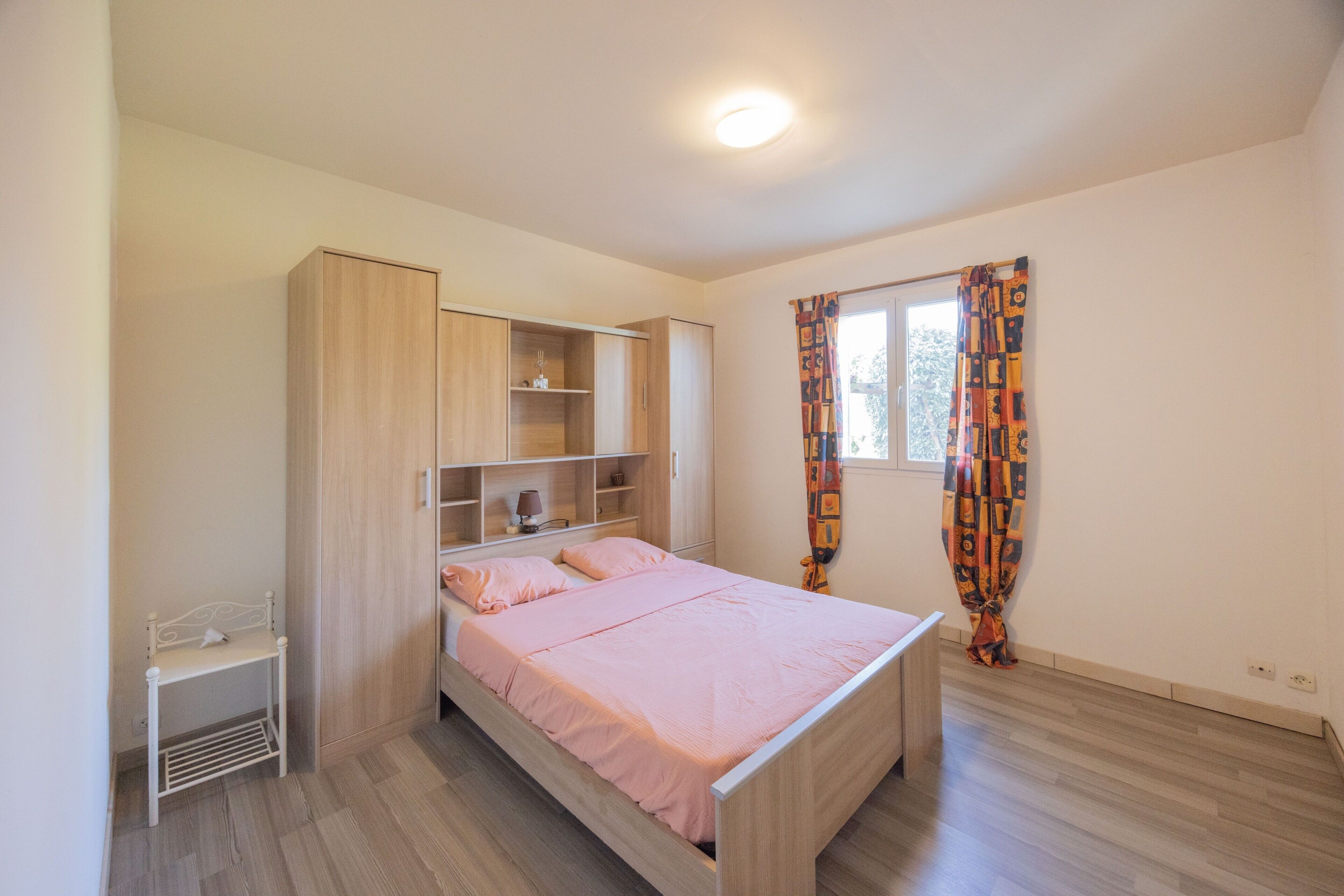 Un dormitorio con cama, armario, estantería, mesita de noche y ventana con cortinas.