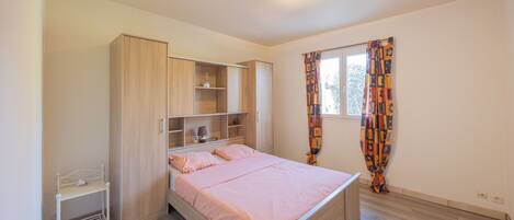Un dormitorio con cama, armario, estantería, mesita de noche y ventana con cortinas.