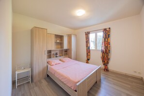 A bedroom with a bed, a wardrobe, a shelf unit, a bedside table, and a window with curtains.