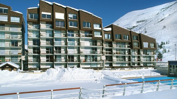 Ein mehrstöckiges Hotel mit Balkonen, einem Swimmingpool und einem verschneiten Berg im Hintergrund.
