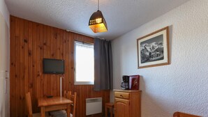 Ein Zimmer mit Schreibtisch aus Holz, einem Sessel, einem Fernseher, einer Heizung, einem Bett mit roter Decke, einem gerahmten Bild an der Wand und einem Fenster mit Vorhängen.