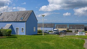 A blue building with a sloped roof, a parking area with cars, and a clear view of the sea in the background. - Brittany Flat, Beach & Pool (Saint-Nic)