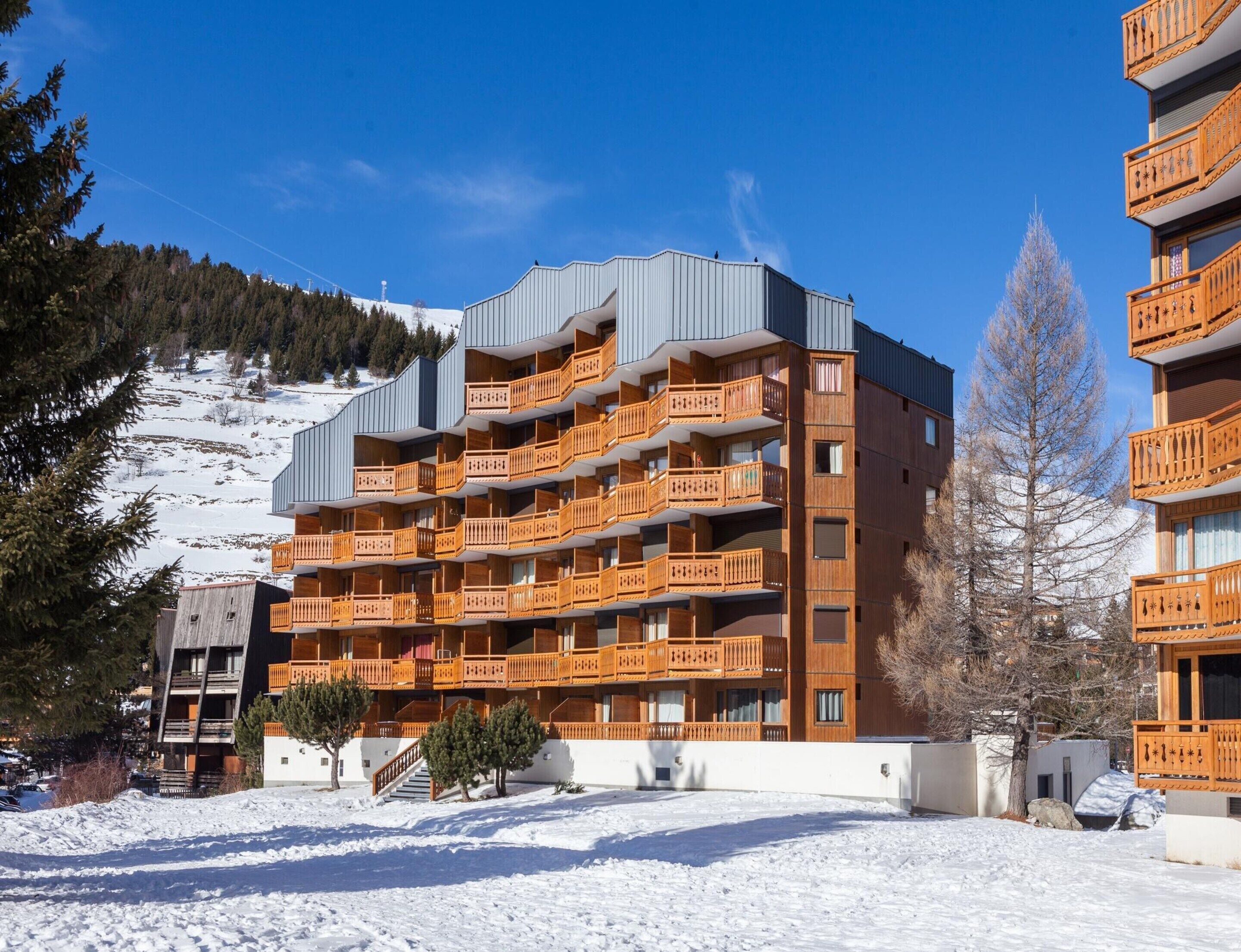 Un hotel de varios pisos con balcones, una pista de esquí al fondo y el suelo cubierto de nieve.