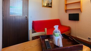 Ein Zimmer mit Holztür, einem roten Sofa, einem kleinen Tisch mit Fernbedienung, einer Toilettenartikeln und einem gerahmten Bild an der Wand sowie einem Regal mit einem Fernseher.