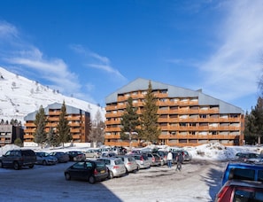Un hotel de varios pisos con balcones, un estacionamiento con coches y una montaña nevada al fondo.