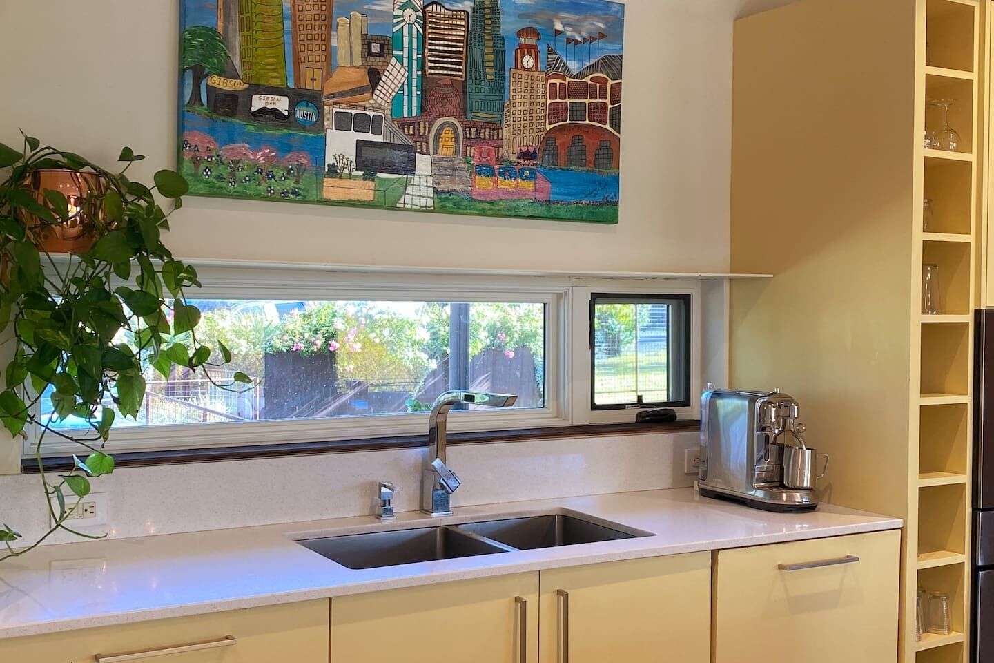 Una cocina con ventana, un lavamanos, una cafetera y un cuadro sobre la ventana.