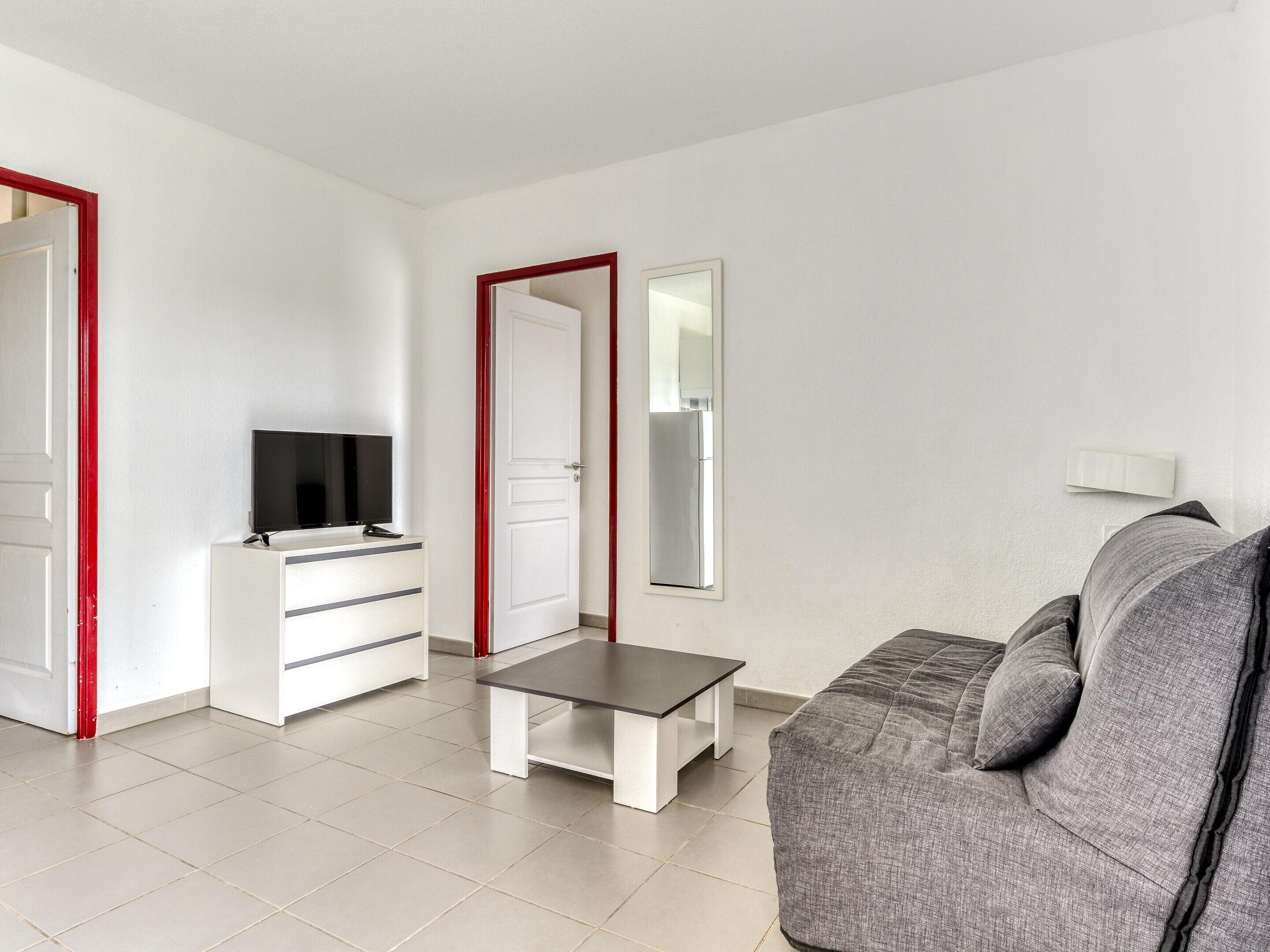 A modern living room with a grey sofa, a small table, a TV on a white cabinet, and a mirror on the wall.