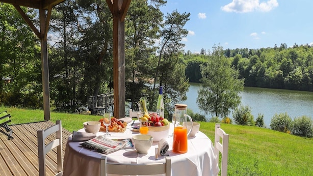 A table set outdoors with breakfast items, a newspaper, and a view of a lake. - 6-person Wooden Chalet in Correze (Meyrignac-l'Eglise)