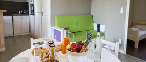 Een ontbijttafel met een fruitschaal, brood en drankjes, een groene bank en een keuken op de achtergrond.