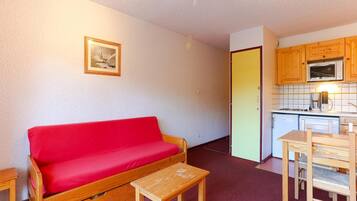 A compact living space with a red sofa, a small wooden table, and a kitchenette with wooden cabinets and a microwave.