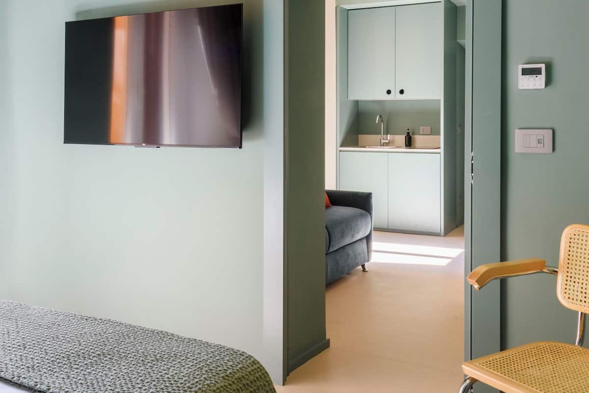 Une chambre d’hôtel avec un lit, une chaise, un téléviseur fixé au mur, une kitchenette et une salle de bain visible à travers une porte ouverte.