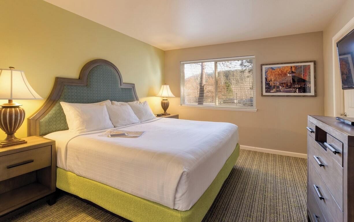 A hotel room with a bed, two bedside lamps, a dresser, a window with blinds, and a framed picture on the wall.