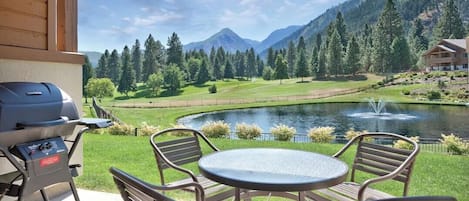 A patio with a grill, a round table, and chairs, overlooking a lake and mountains.