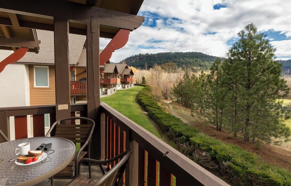 A balcony with a table, chairs, and a view of a forested area and buildings in the background.
