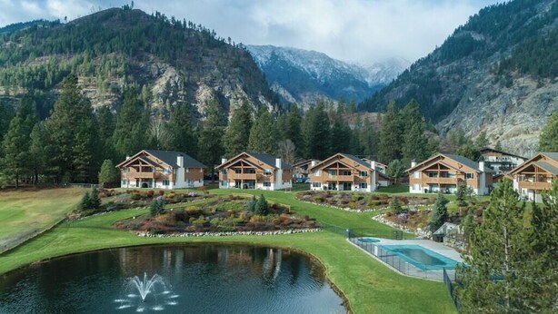 Un resort con varias cabañas de madera, un estanque con una fuente y montañas al fondo.