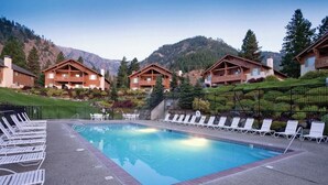 Un área de piscina con sillones de descanso, cabañas de madera y montañas al fondo.