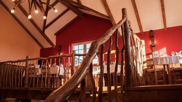 Ein rustikales Interieur mit freiliegenden Holzbalken, einer roten Wand und einem Essbereich mit Tischen und Stühlen.