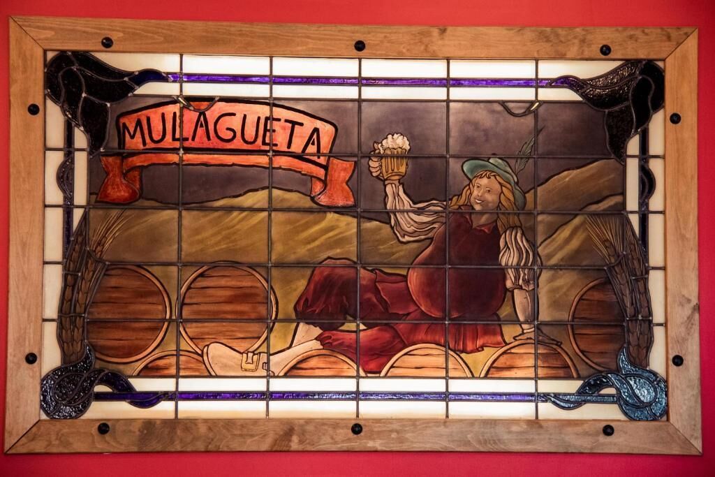 Una ventana de vitral con la palabra 'MULAGUETA' y una figura sosteniendo una jarra.