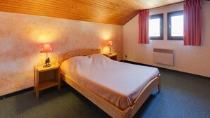 A bedroom with a wooden ceiling, two bedside tables with lamps, a bed, a window with curtains, and an air conditioning unit. - Bright Apartment Close to Ski-slopes (Les Deux Alpes)