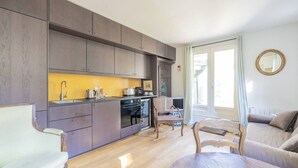 Une cuisine moderne dotée d’appareils électroménagers intégrés, un coin repas avec une table et des chaises en bois, et un salon avec un canapé et un fauteuil.