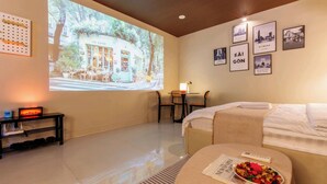 Ein Hotelzimmer mit einem Bett, einem kleinen Tisch mit Fernbedienung, einem projizierten Bild eines Cafés und gerahmten Bildern an der Wand.