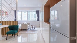 Ein modernes Hotelzimmer mit einem weißen Kühlschrank, einem Esstisch, einem petrolfarbenen Sessel, einer Couch und Blick auf die Stadt durch große Fenster.