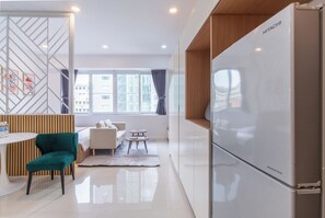 A modern hotel room with a white refrigerator, a dining table, a teal chair, a sofa, and a view of the city through large windows. - Le Places Old Condos Central Nguyen Hue  (Ho Chi Minh City)