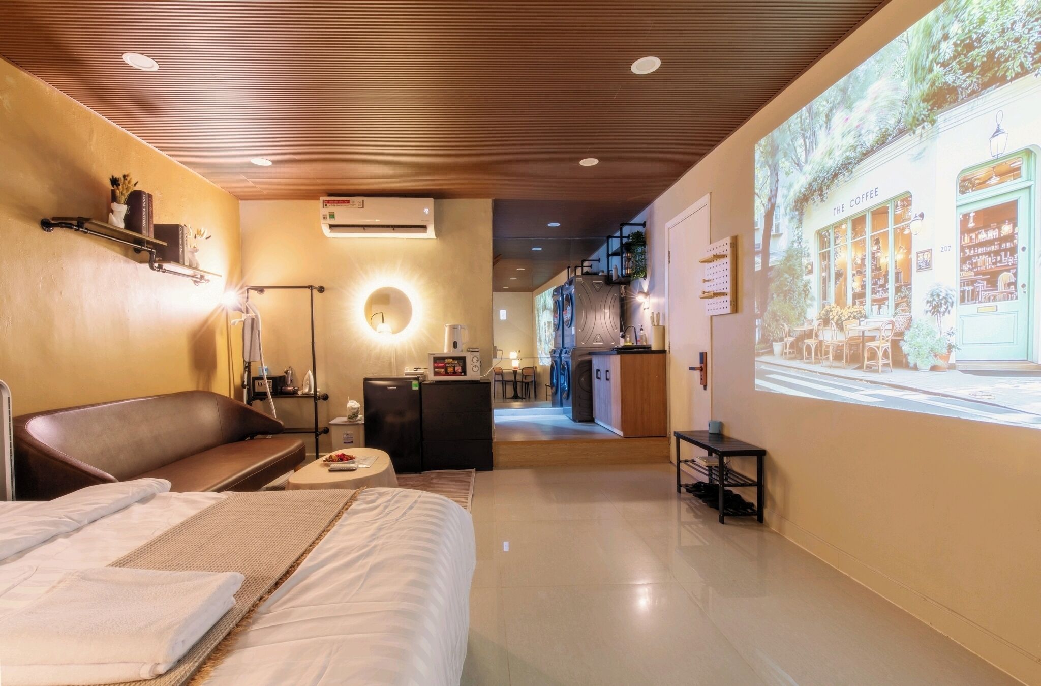 A hotel room with a bed, a seating area, a kitchenette, and a projection of a coffee shop on the wall.