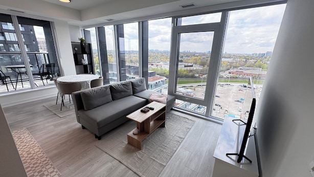 En moderne stue med en grå sofa, et lille træbord og et stort vindue med udsigt over byen.