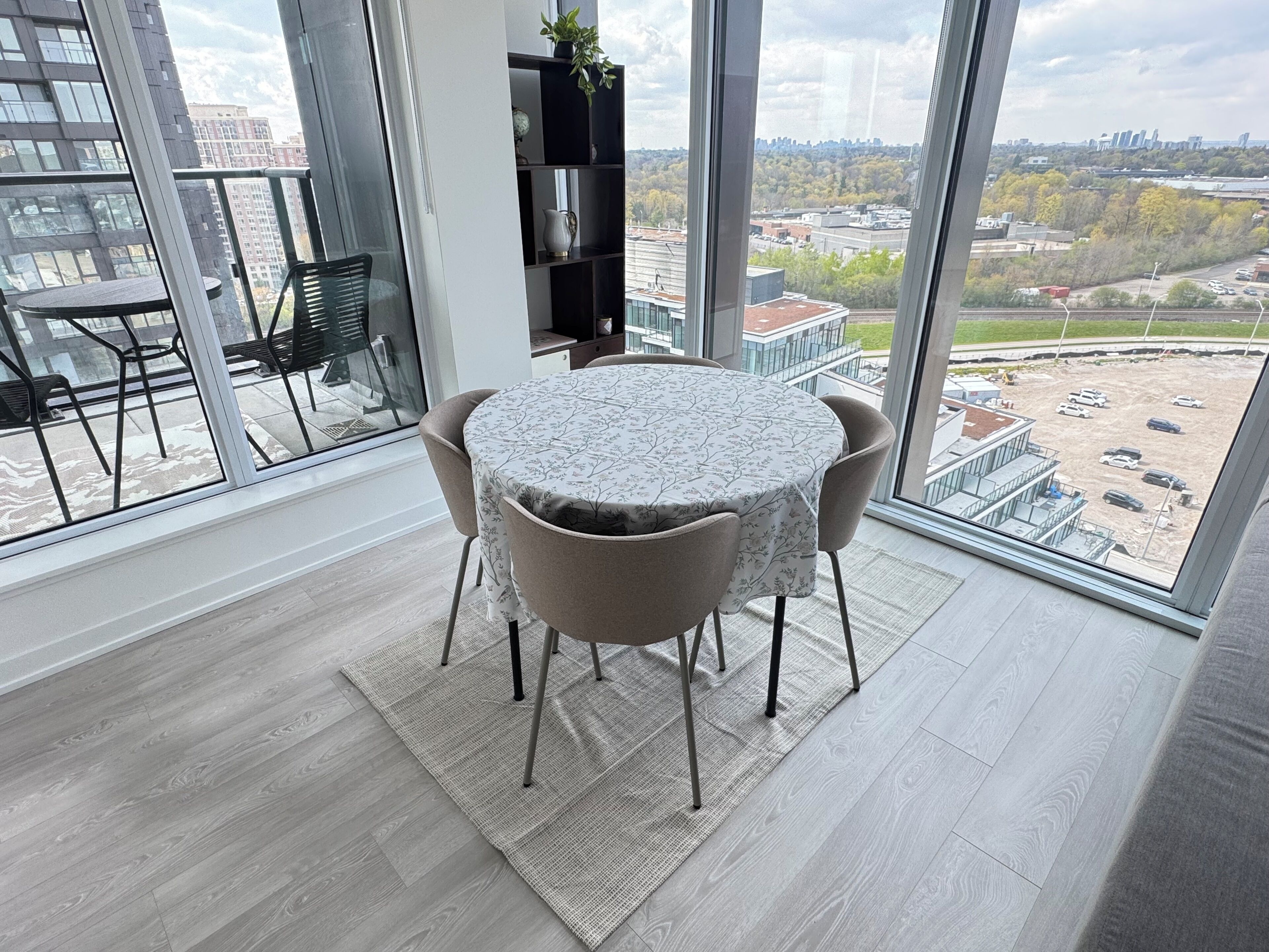 A modern dining area with a round table and chairs, a bookshelf, and a large window with a city view.