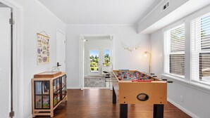 Een kamer met een tafelvoetbaltafel, een kast met glazen deuren, een deur naar een balkon en een raam met jaloezieën.