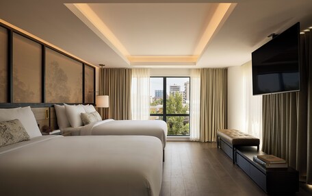 A hotel room with two beds, a TV, a window with curtains, and a view of the city.