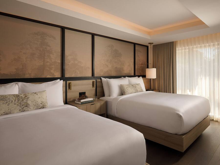 Habitación de hotel con dos camas, una mesita de noche, una lámpara y un ventanal con cortinas.