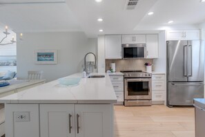 A modern kitchen with white cabinetry, stainless steel appliances, a large island with a bowl, and a painting on the wall. - Carlos Pointe #126 - Endless Beach (Fort Myers Beach)