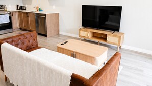 A modern living room with a sofa, a coffee table, a TV, and a kitchen area with cabinets and appliances.