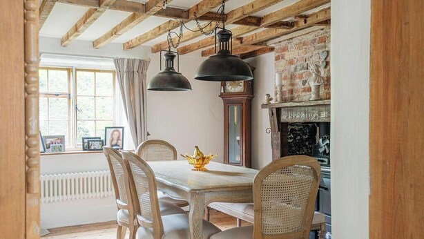 Une salle à manger avec une table en bois, des chaises en osier, un lustre, une horloge et une cheminée.