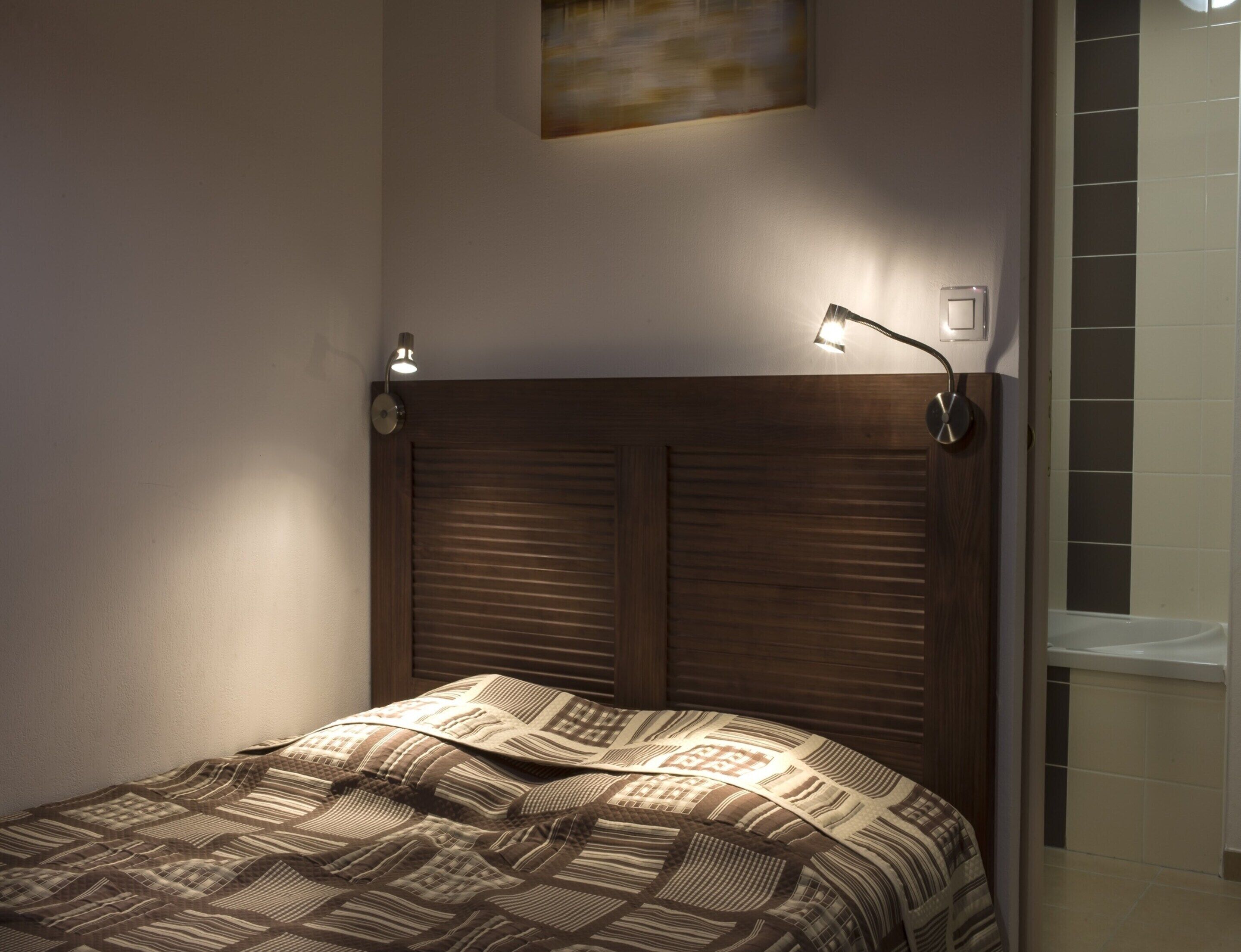 Une chambre à coucher avec une tête de lit en bois, une couvre-lit à motifs, deux lampes de chevet et un luminaire fixé au mur.