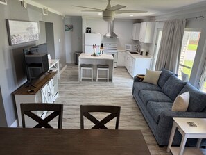 Eine moderne Küche mit weißen Schränken, ein Essbereich mit Holztisch und -stühlen sowie ein graues Sofa mit Blick ins Freie.