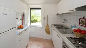 Eine kleine Küche mit weißen Schränken, einem Einbauofen, einer Spüle und einem Fenster mit Blick auf Grünflächen.
