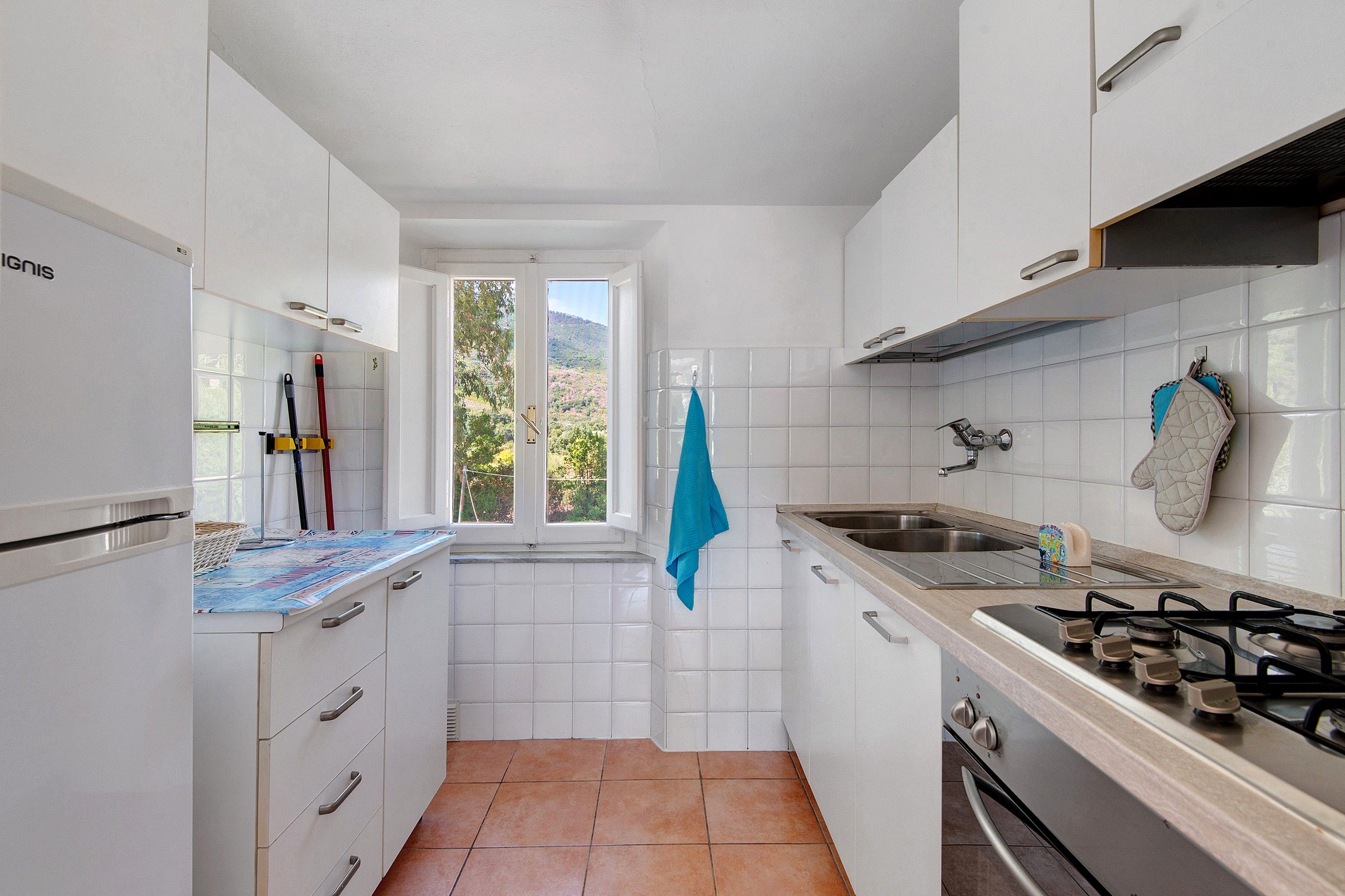 Cocina compacta con armarios blancos, estufa a gas, lavamanos y ventana con vistas a zonas verdes.
